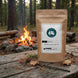 Kraft paper tea bag with a label on a wooden table in a forest setting with a campfire.
